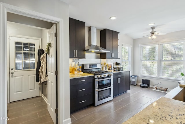 kitchen featuring range with two ovens, ceiling fan, light stone countertops, wall chimney range hood, and backsplash