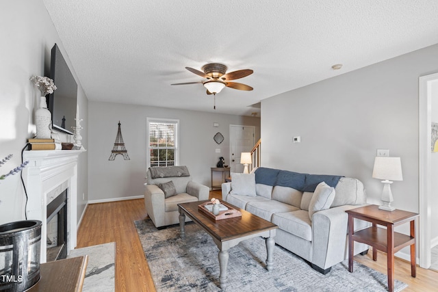 living area with ceiling fan, a textured ceiling, baseboards, light wood finished floors, and a tiled fireplace