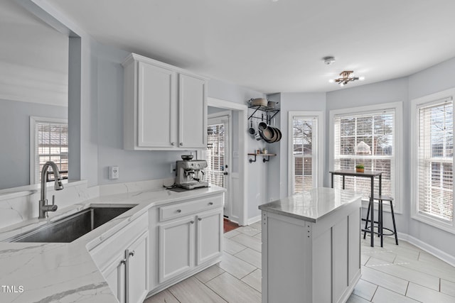 kitchen featuring light stone counters, white cabinets, a sink, a kitchen island, and baseboards