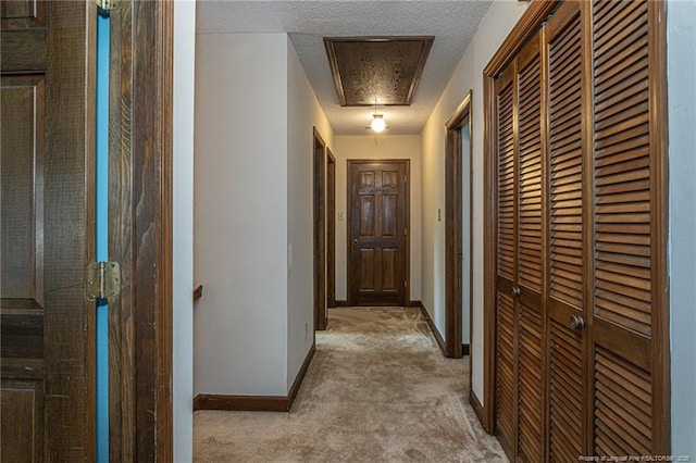 corridor featuring attic access, baseboards, a textured ceiling, and light colored carpet