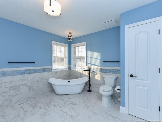 bathroom featuring marble finish floor, visible vents, a freestanding bath, toilet, and wainscoting