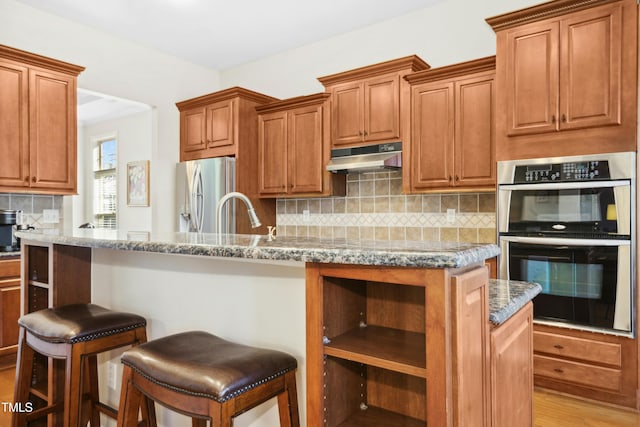 kitchen featuring tasteful backsplash, a kitchen island with sink, stainless steel appliances, under cabinet range hood, and a sink