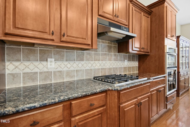 kitchen with brown cabinetry, appliances with stainless steel finishes, dark stone countertops, under cabinet range hood, and backsplash