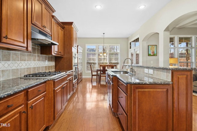 kitchen with a kitchen island with sink, under cabinet range hood, a sink, appliances with stainless steel finishes, and light wood-type flooring