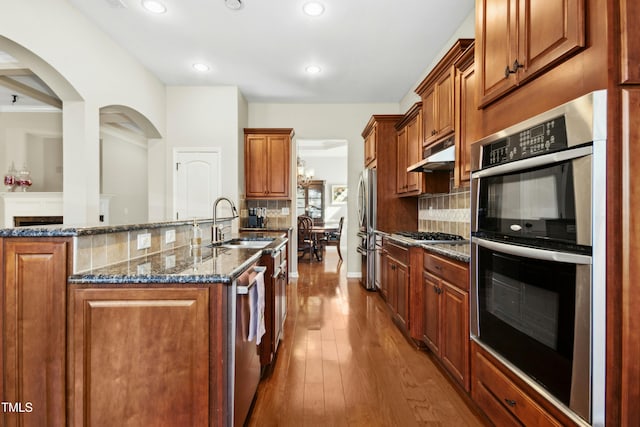 kitchen with under cabinet range hood, dark wood-style flooring, a sink, appliances with stainless steel finishes, and dark stone counters