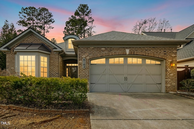 french country home featuring a shingled roof, concrete driveway, brick siding, and an attached garage