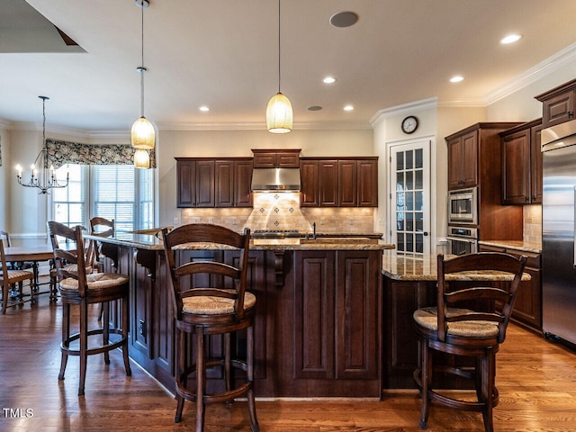 kitchen with dark brown cabinetry, light stone counters, dark wood-style flooring, built in appliances, and under cabinet range hood