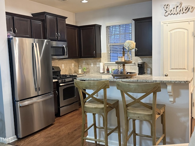 kitchen featuring dark brown cabinetry, decorative backsplash, dark wood-style floors, a kitchen breakfast bar, and stainless steel appliances