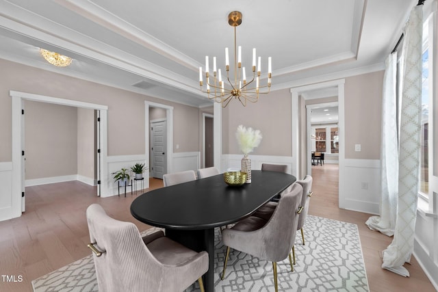 dining space featuring a tray ceiling, crown molding, a notable chandelier, light wood finished floors, and wainscoting