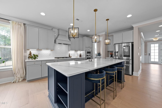 kitchen featuring custom range hood, appliances with stainless steel finishes, hanging light fixtures, gray cabinets, and a large island with sink