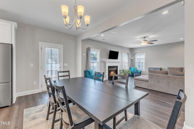 dining space with recessed lighting, ceiling fan with notable chandelier, wood finished floors, baseboards, and a lit fireplace