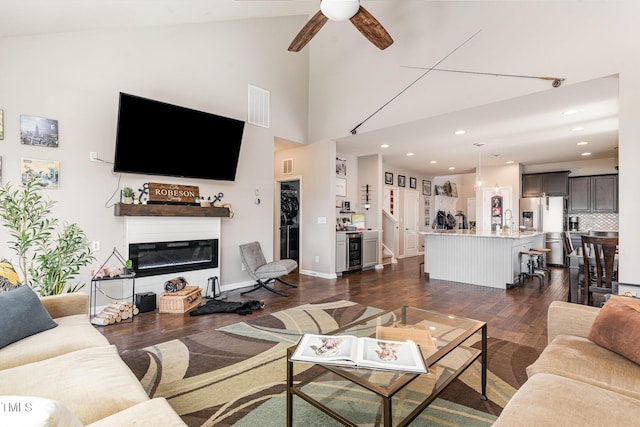 living room with visible vents, dark wood finished floors, a glass covered fireplace, ceiling fan, and high vaulted ceiling