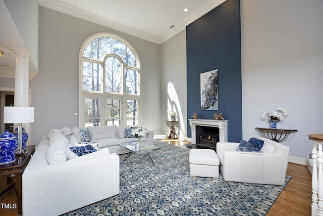 living area with wood finished floors, a lit fireplace, crown molding, a towering ceiling, and ornate columns