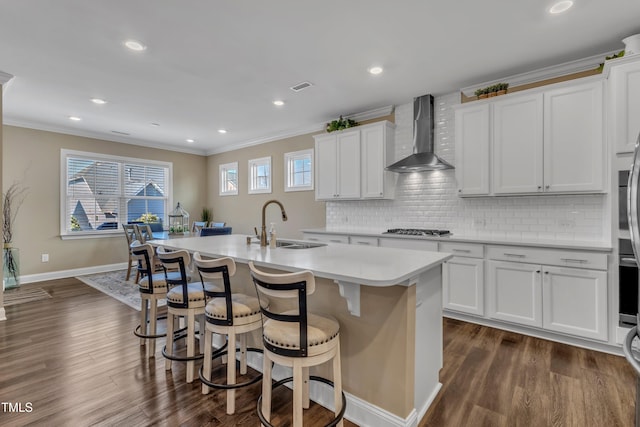 kitchen with a center island with sink, ornamental molding, a sink, stainless steel gas stovetop, and wall chimney exhaust hood