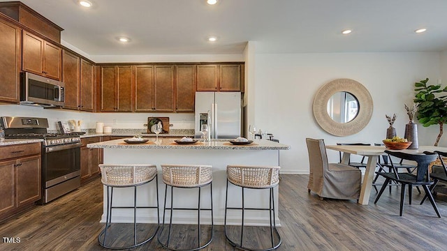 kitchen with dark wood finished floors, recessed lighting, appliances with stainless steel finishes, a kitchen island with sink, and light stone countertops