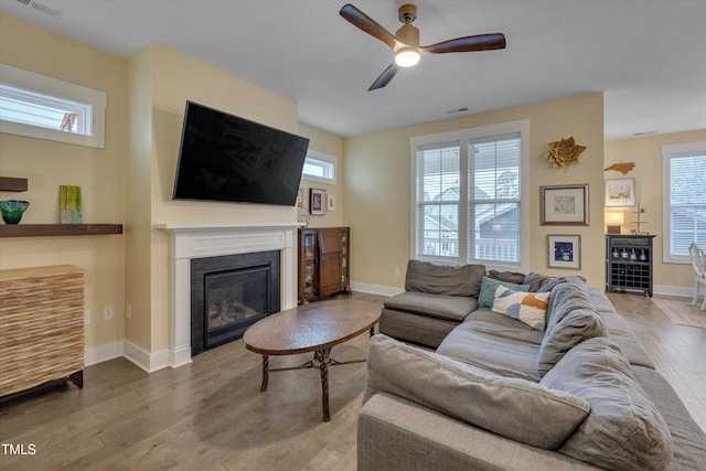 living area featuring a ceiling fan, a glass covered fireplace, baseboards, and wood finished floors