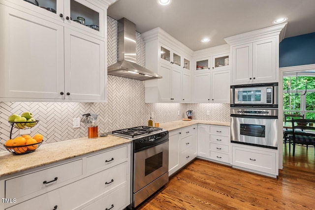kitchen with appliances with stainless steel finishes, wall chimney exhaust hood, white cabinets, light wood-style flooring, and decorative backsplash
