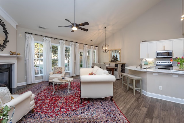 living room featuring ceiling fan with notable chandelier, dark wood-type flooring, visible vents, baseboards, and a glass covered fireplace