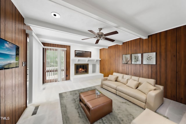 living area featuring beamed ceiling, ceiling fan, wooden walls, and a glass covered fireplace