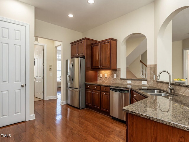 kitchen with stainless steel appliances, a sink, dark wood-style floors, decorative backsplash, and dark stone counters