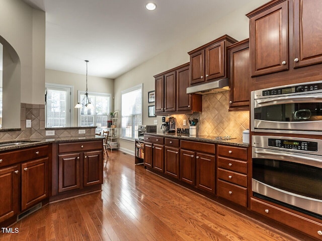 kitchen with black electric stovetop, stainless steel double oven, under cabinet range hood, dark wood-style floors, and dark stone countertops