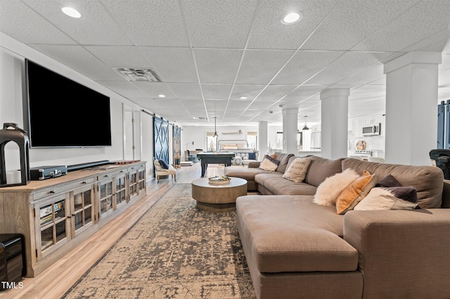 living area with light wood-type flooring, visible vents, a drop ceiling, and ornate columns