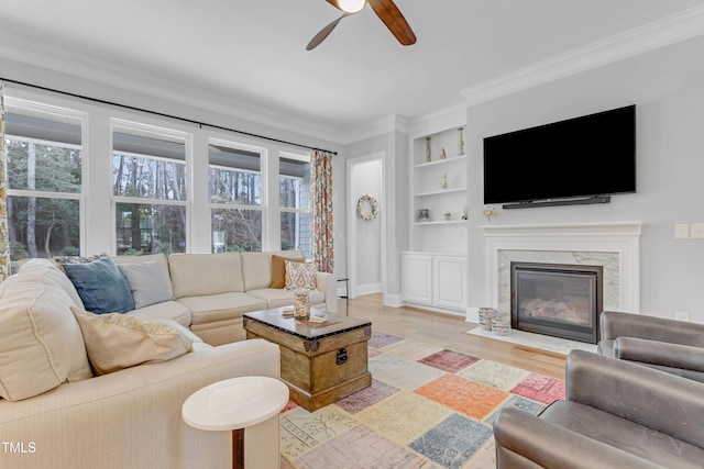 living room featuring built in shelves, a premium fireplace, a ceiling fan, light wood finished floors, and crown molding