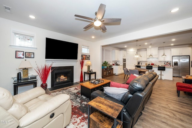 living room with light wood finished floors, visible vents, a fireplace with flush hearth, ceiling fan, and recessed lighting