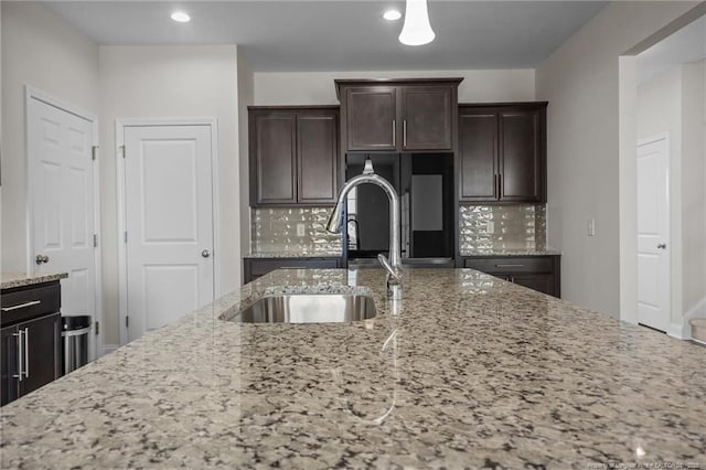 kitchen featuring light stone counters, refrigerator, backsplash, a sink, and dark brown cabinetry