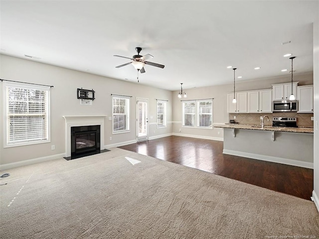 unfurnished living room featuring baseboards, a fireplace with flush hearth, ceiling fan, dark colored carpet, and a sink