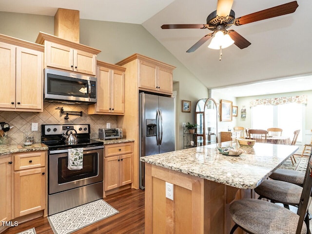 kitchen with dark wood-style floors, stainless steel appliances, light brown cabinetry, vaulted ceiling, and a kitchen bar