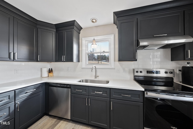 kitchen with light wood finished floors, appliances with stainless steel finishes, light countertops, wall chimney range hood, and a sink