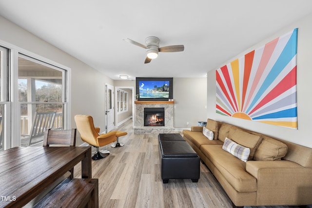 living area featuring a ceiling fan, a stone fireplace, light wood-style flooring, and baseboards