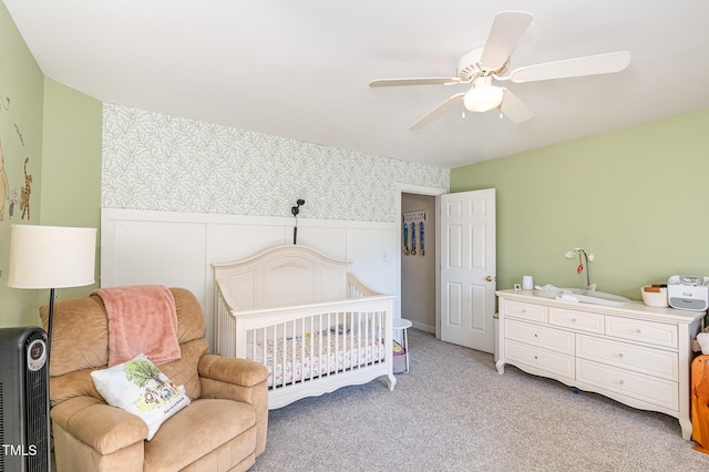 bedroom with light carpet, wallpapered walls, a crib, a ceiling fan, and a wainscoted wall