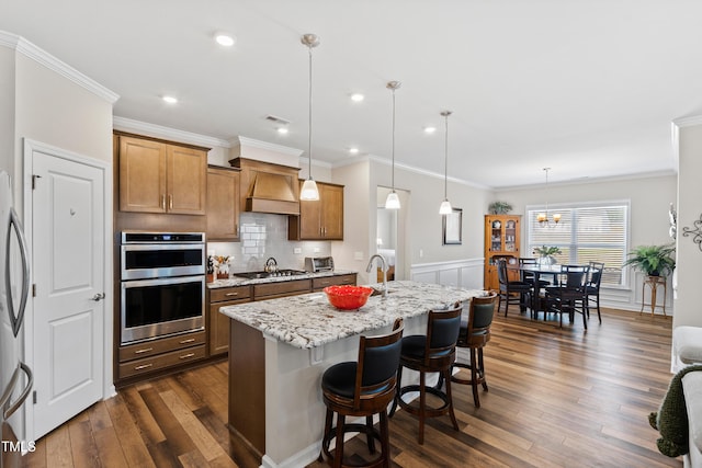 kitchen featuring dark wood-style floors, custom range hood, brown cabinetry, appliances with stainless steel finishes, and a kitchen island with sink