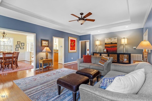 living area with crown molding, wood-type flooring, a raised ceiling, a ceiling fan, and baseboards