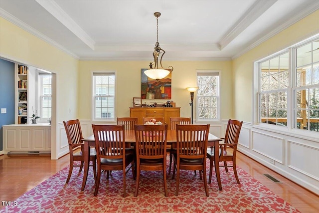 dining space featuring light wood finished floors, visible vents, a raised ceiling, wainscoting, and crown molding