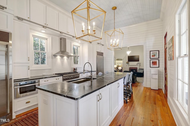 kitchen featuring an island with sink, wall chimney exhaust hood, high quality appliances, white cabinetry, and a sink