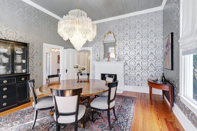 dining space with wallpapered walls, baseboards, a fireplace with raised hearth, ornamental molding, and wood finished floors