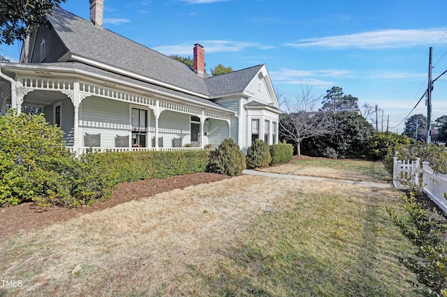 view of side of property featuring a shingled roof, a lawn, a chimney, covered porch, and fence