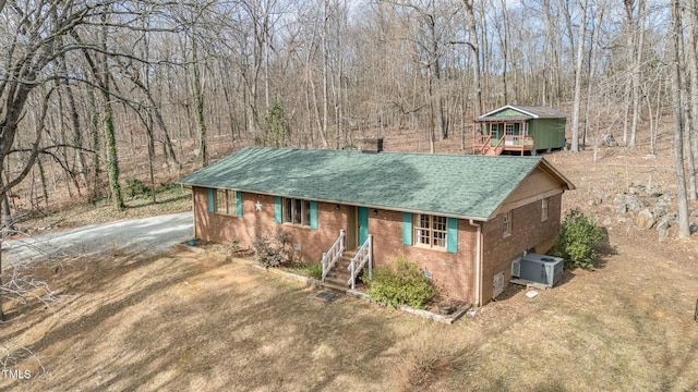 ranch-style house with central AC, brick siding, a shingled roof, driveway, and a wooded view