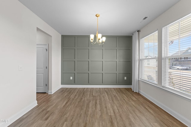 unfurnished dining area featuring a decorative wall, wood finished floors, visible vents, baseboards, and an inviting chandelier