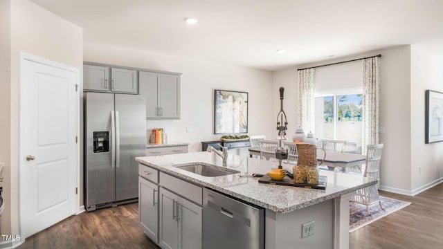 kitchen with light stone counters, stainless steel appliances, gray cabinetry, a sink, and an island with sink