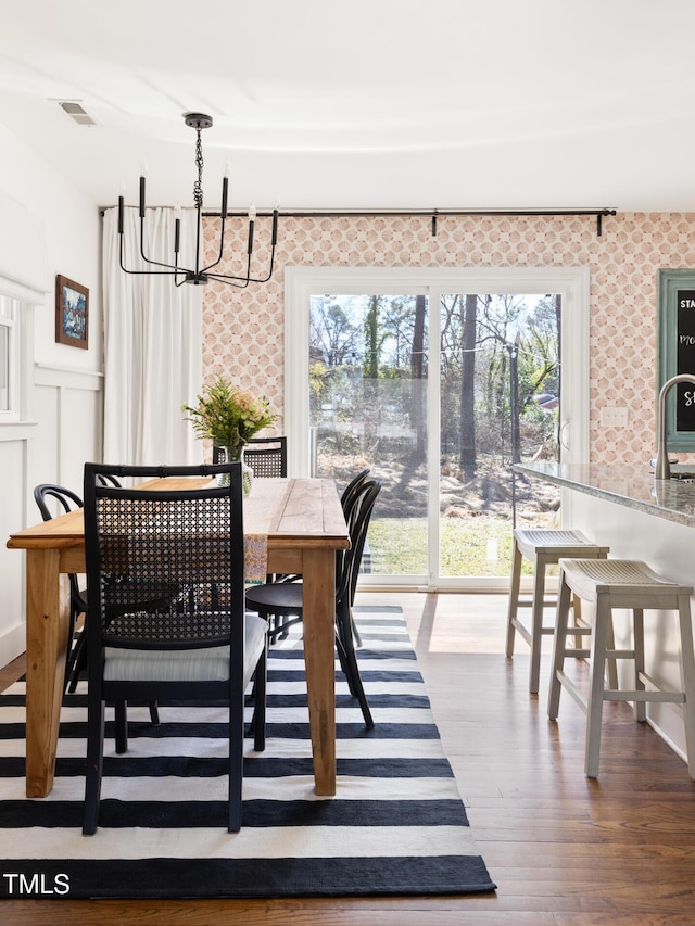 dining room featuring a notable chandelier, wood finished floors, visible vents, wainscoting, and wallpapered walls