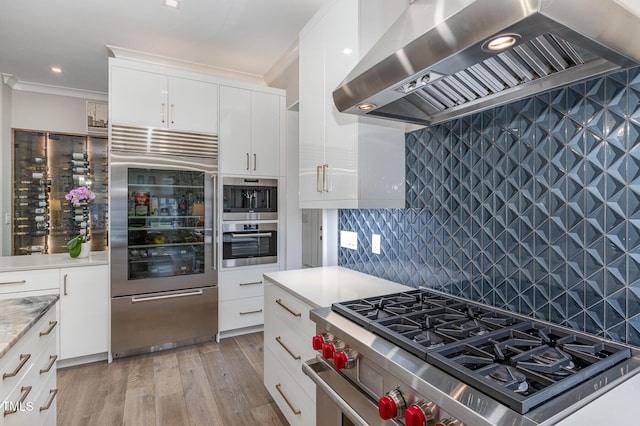 kitchen featuring high end appliances, wall chimney exhaust hood, light countertops, light wood-type flooring, and backsplash