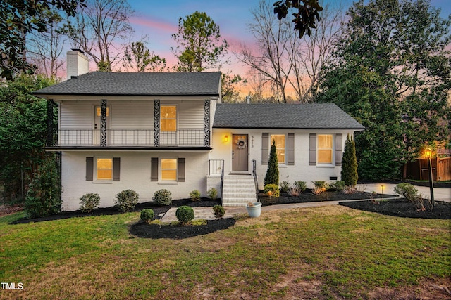 split level home featuring brick siding, a chimney, a shingled roof, a balcony, and a front lawn