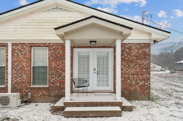 snow covered property entrance featuring french doors, ac unit, and brick siding