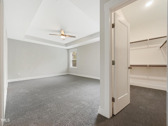 empty room featuring a tray ceiling, dark colored carpet, a ceiling fan, and baseboards