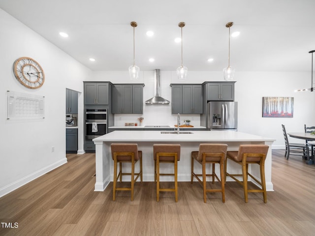 kitchen featuring light wood finished floors, a center island with sink, wall chimney exhaust hood, stainless steel appliances, and light countertops