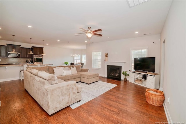 living area featuring recessed lighting, ceiling fan with notable chandelier, a fireplace with flush hearth, baseboards, and dark wood finished floors